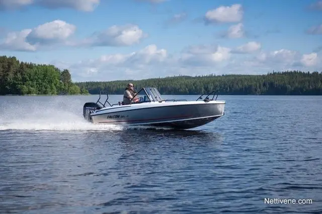 A man drives a Falcon boat across a calm lake, creating waves, with trees and blue skies in the background.